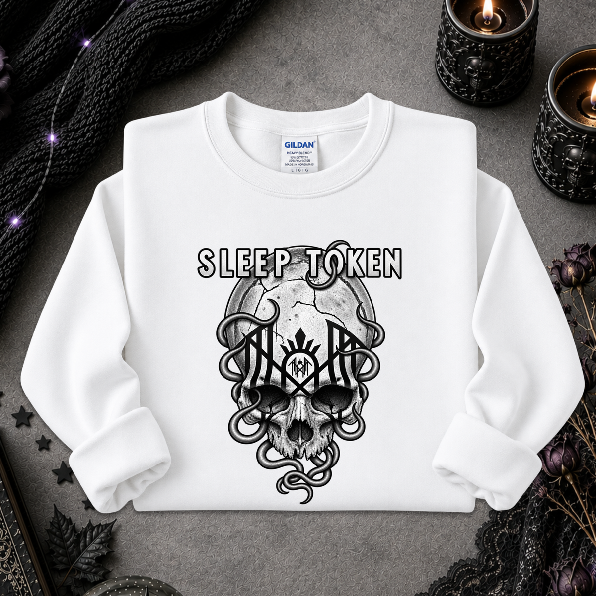 A white long-sleeve crewneck sweatshirt with a black graphic print of a skull, snakes, and the text 'SLEEP TOKEN' on the front. The sweatshirt is laid flat on a dark gray textured surface surrounded by gothic props including black lace, dried flowers, and lit candles.