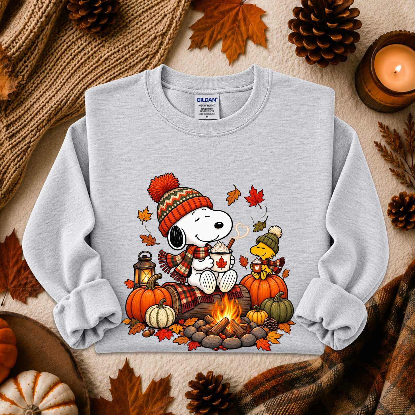 A light gray long-sleeve sweatshirt featuring a colorful autumn-themed graphic of Snoopy and Woodstock by a campfire, surrounded by pumpkins, fall leaves, pinecones, a candle, and cozy knit textiles in a styled flat-lay arrangement.