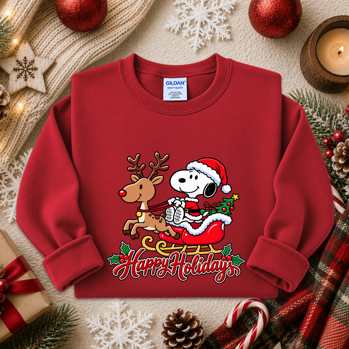 A red long-sleeve crewneck sweatshirt featuring a cartoon Snoopy and Woodstock in Santa suits with a Christmas tree and 'Happy Holidays' text, styled in a cozy holiday flat-lay with pine branches, ornaments, a candle, and a gift box.