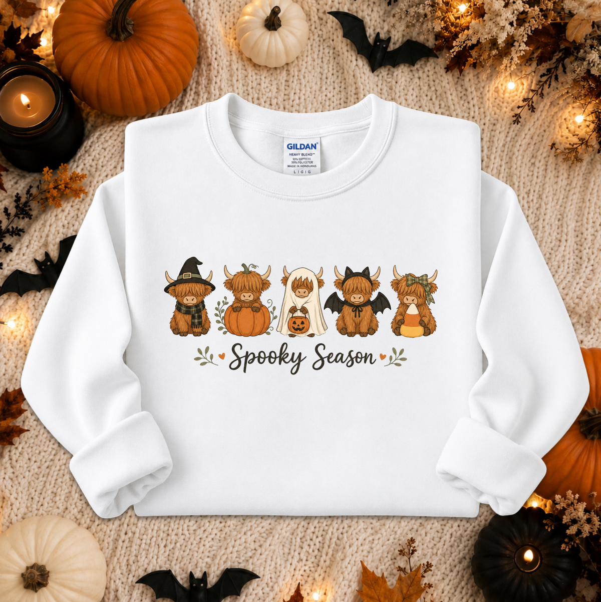 A white long-sleeve crewneck sweatshirt laid flat on a beige knit blanket, surrounded by Halloween decorations including pumpkins, small black bats, leaves, and warm string lights. The sweatshirt has a printed graphic of five cartoon bogarts with the words 'Spooky Season' underneath.