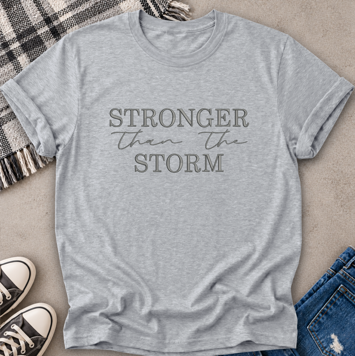 A light heather gray short-sleeve t-shirt with the text 'STRONGER than the STORM' printed on the front in a dark gray tone. The shirt is styled in a flat-lay arrangement on a textured beige surface, surrounded by a black and white plaid blanket, black and white sneakers, and blue jeans.