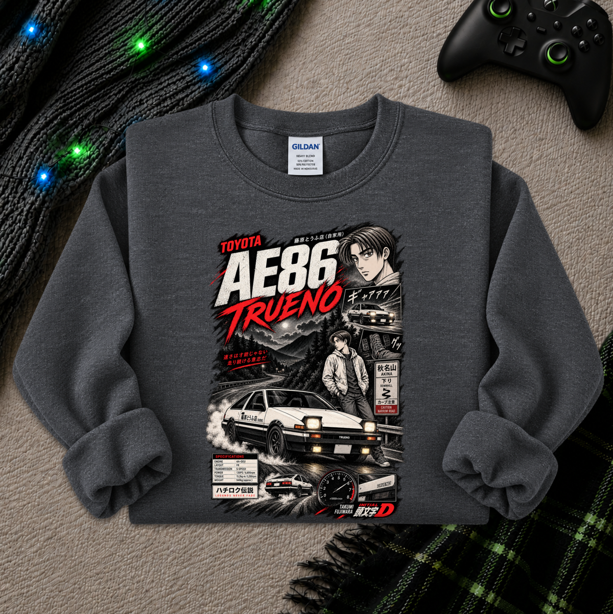 A dark gray crewneck sweatshirt featuring a detailed graphic print of the Toyota AE86 Trueno from the movie Initial D, styled in a cozy flat-lay setting with a black knit sweater, glowing fairy lights, a video game controller, and a green plaid blanket.