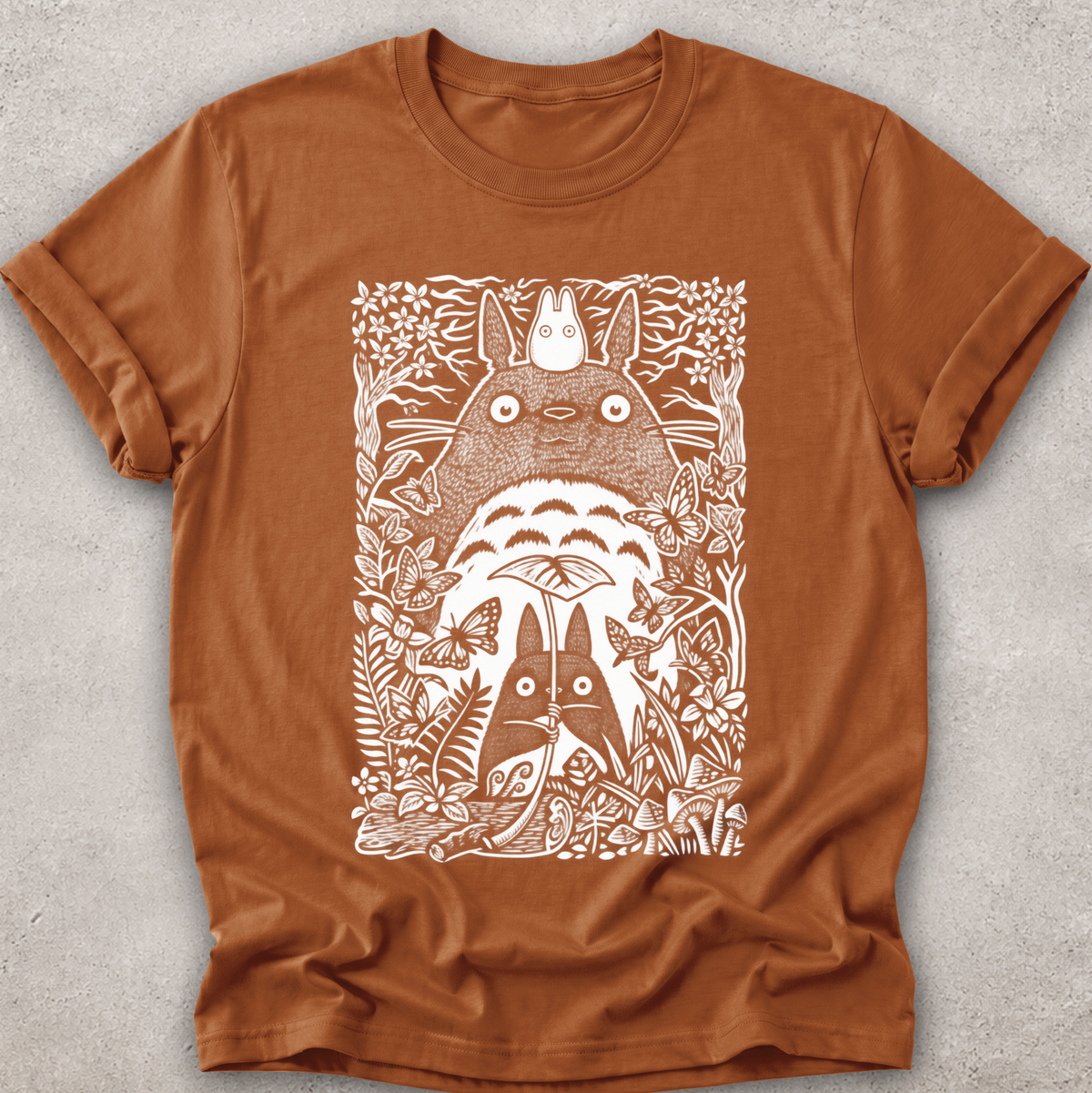 A rust-brown short-sleeve t-shirt with a white screen-printed graphic of Totoro and other My Neighbor Totoro characters surrounded by foliage, laid flat on a textured gray background.