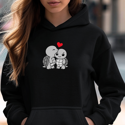 A woman wearing a black pullover hoodie with a cute cartoon graphic of two kissing sea turtles under a small red heart on the chest.