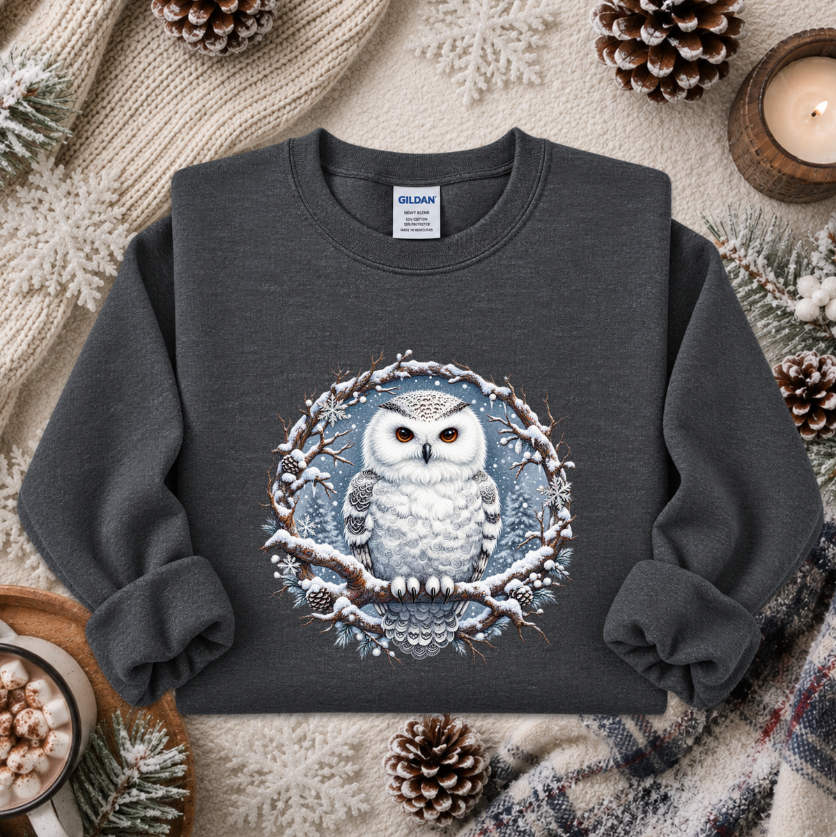 A dark gray crewneck sweatshirt featuring a detailed graphic of a snowy owl in a winter tree circle, styled in a cozy flat-lay with pinecones, a candle, and a knit blanket. The Gildan brand tag is visible inside the collar.