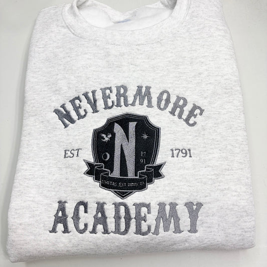 A folded light heather gray sweatshirt featuring a dark gray graphic print that reads 'NEVERMORE ACADEMY' with a crest-style emblem in the center and 'EST 1791' on either side.