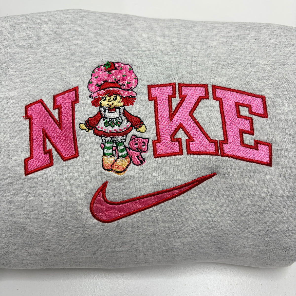 A close-up of a folded heather gray sweatshirt featuring a pink embroidered 'NIKE' design with a strawberry shortcake character and a red swoosh logo.
