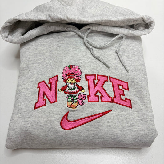 A folded light gray heather hoodie with a pink and red embroidered design on the front. The design features the word 'NIKE' in large pink letters with a cartoon strawberry shortcake character in the center and a pink Nike swoosh below.
