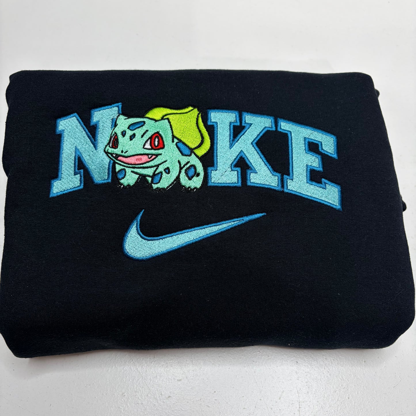 A folded black sweatshirt featuring a light blue embroidered 'NIKE' logo, a Bulbasaur character graphic, and a matching Nike swoosh on the front.