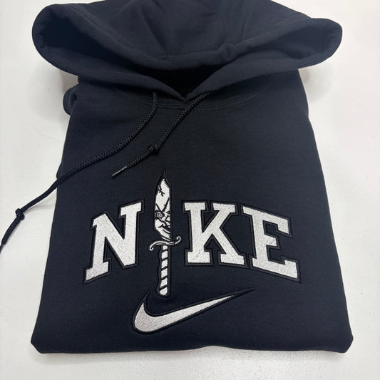A folded black Nike hoodie featuring a custom embroidered design on the front. The design spells 'NIKE' in white varsity-style block letters, with the letter 'I' replaced by a detailed white and black dagger motif, and a white Nike swoosh embroidered underneath.