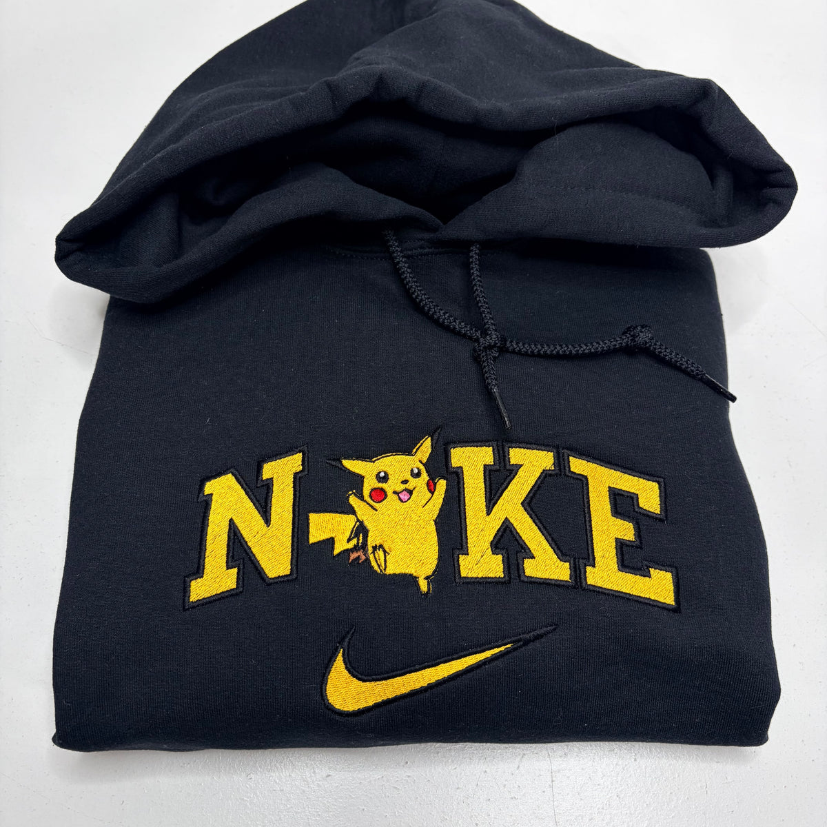 A black hoodie featuring a custom 'Nike design with Pikachu embroidered in yellow and black, and a yellow Nike swoosh below, shown on a white background.