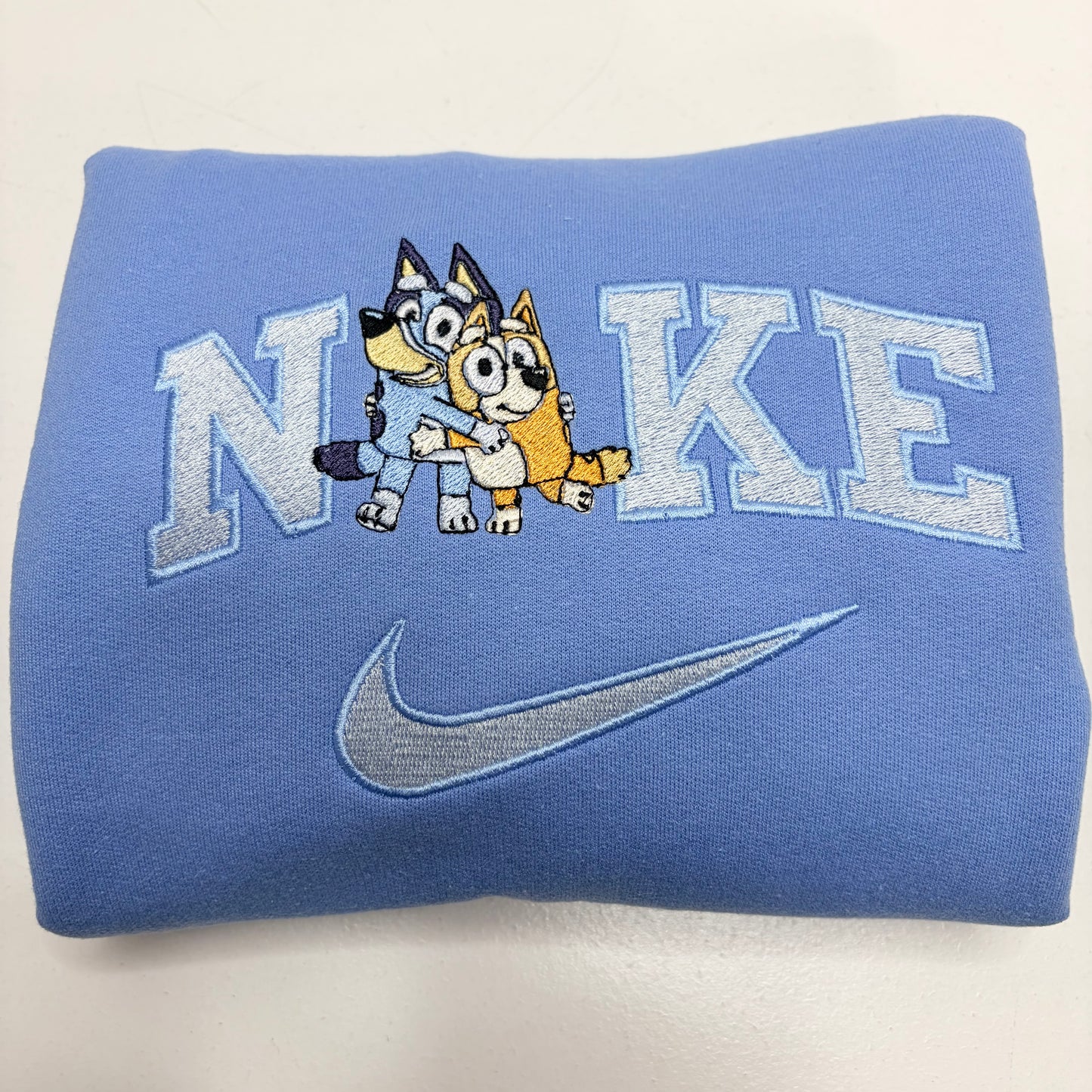 A folded light blue sweatshirt featuring an embroidered design with the word 'NiKE' in light blue varsity letters, a cartoon bluey and bingo dog characters in the center, and a gray embroidered Nike swoosh below.