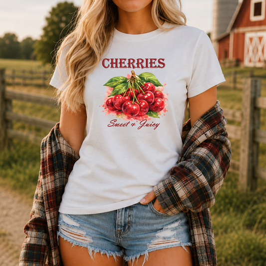 A woman wearing a white graphic t-shirt with a red and green cherry print that reads 'CHERRIES Sweet & Juicy', paired with a plaid flannel shirt and denim shorts, set against a rustic outdoor background with a red barn.