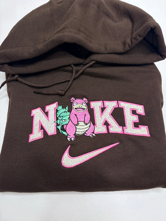 A brown pullover hoodie featuring a pink and white embroidered 'NIKE' graphic with a pink character and a pink swoosh on the front.