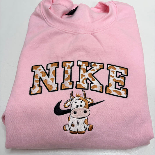 A folded light pink sweatshirt featuring a large embroidered 'NIKE' logo in a cow-print pattern with black outlines, a black Nike swoosh, and a cartoon cow graphic below.