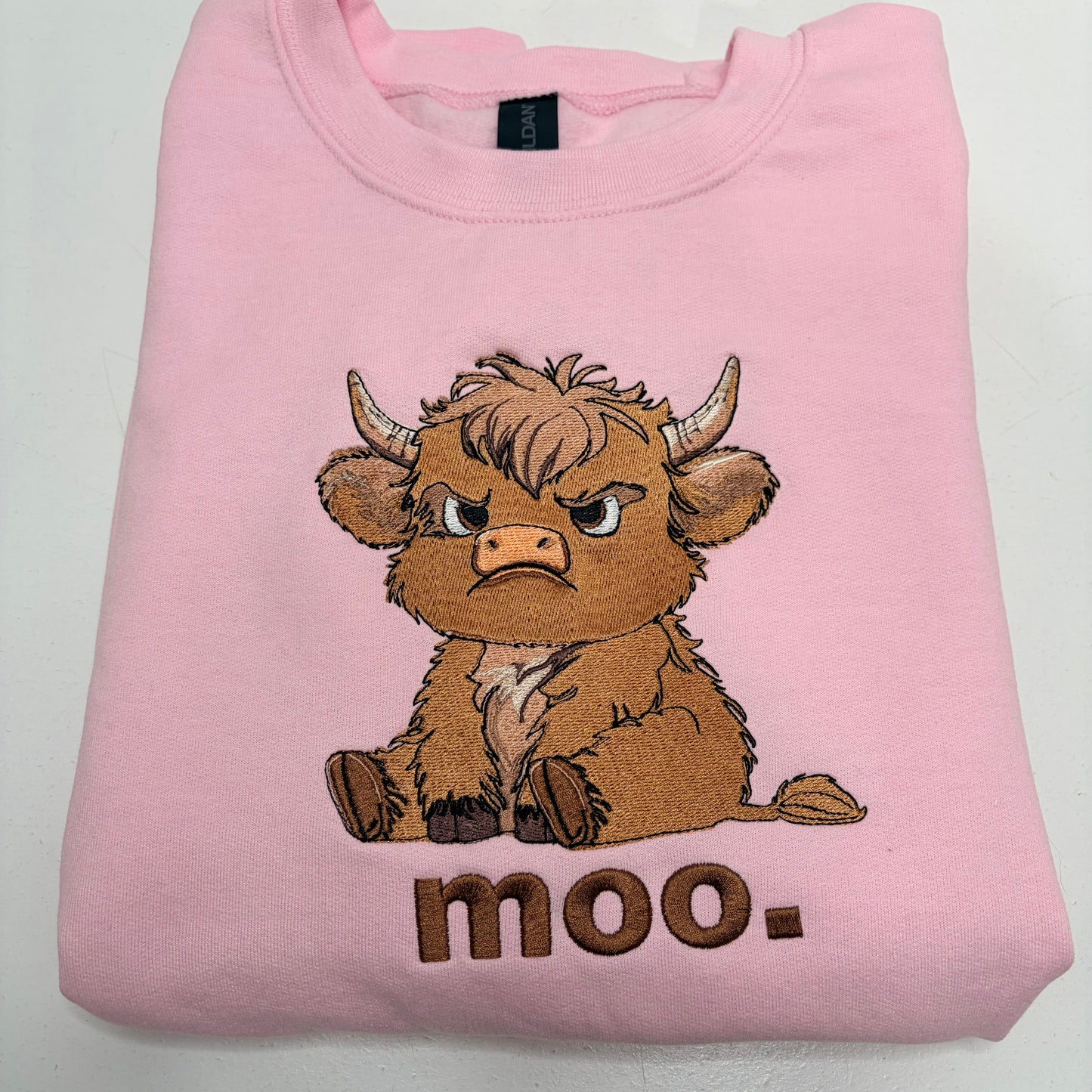 A folded light pink sweatshirt featuring a cartoon illustration of an angry brown Highland cow with the word 'moo.' embroidered below it, set against a plain white background.
