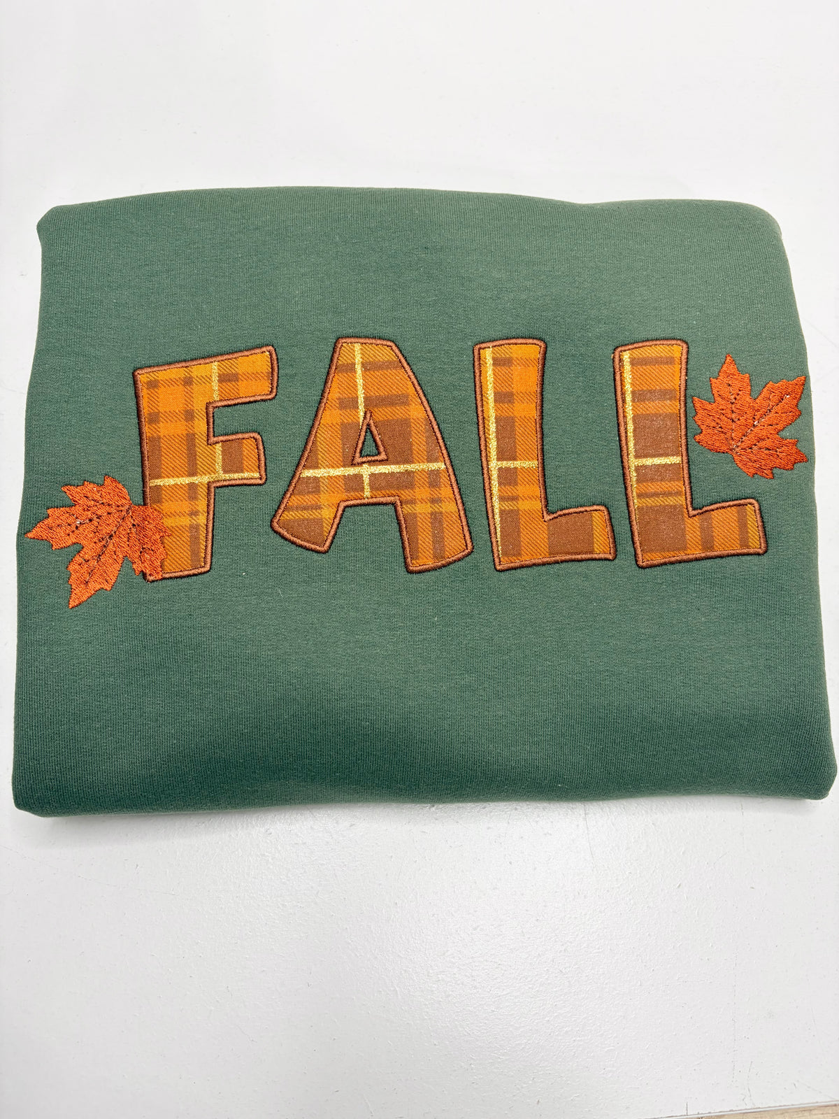 A folded green sweatshirt featuring the word 'FALL' in orange and brown plaid letters, accented with two orange maple leaves on either side, displayed on a white background.