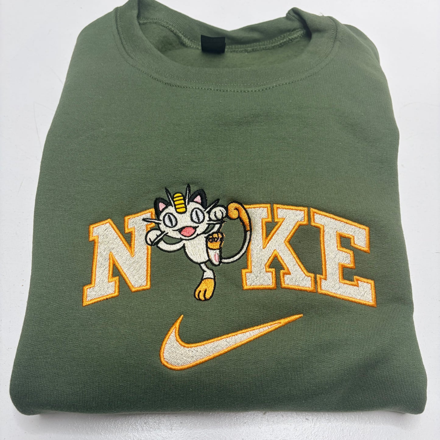 A folded olive green sweatshirt featuring a large embroidered 'NIKE' logo with a white and yellow cartoon meowth pokemon character, and a matching embroidered Nike swoosh below.