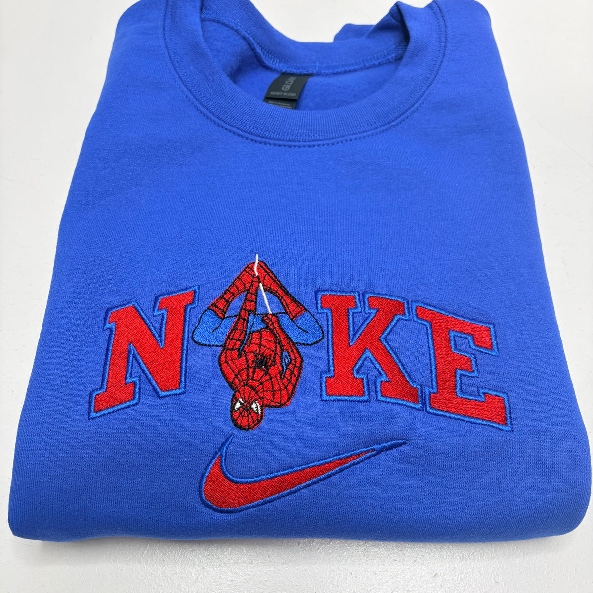 A folded royal blue sweatshirt featuring a red and blue embroidered design that spells 'NOKE' with a Spider-Man figure inside the 'O', and a red Nike swoosh below.
