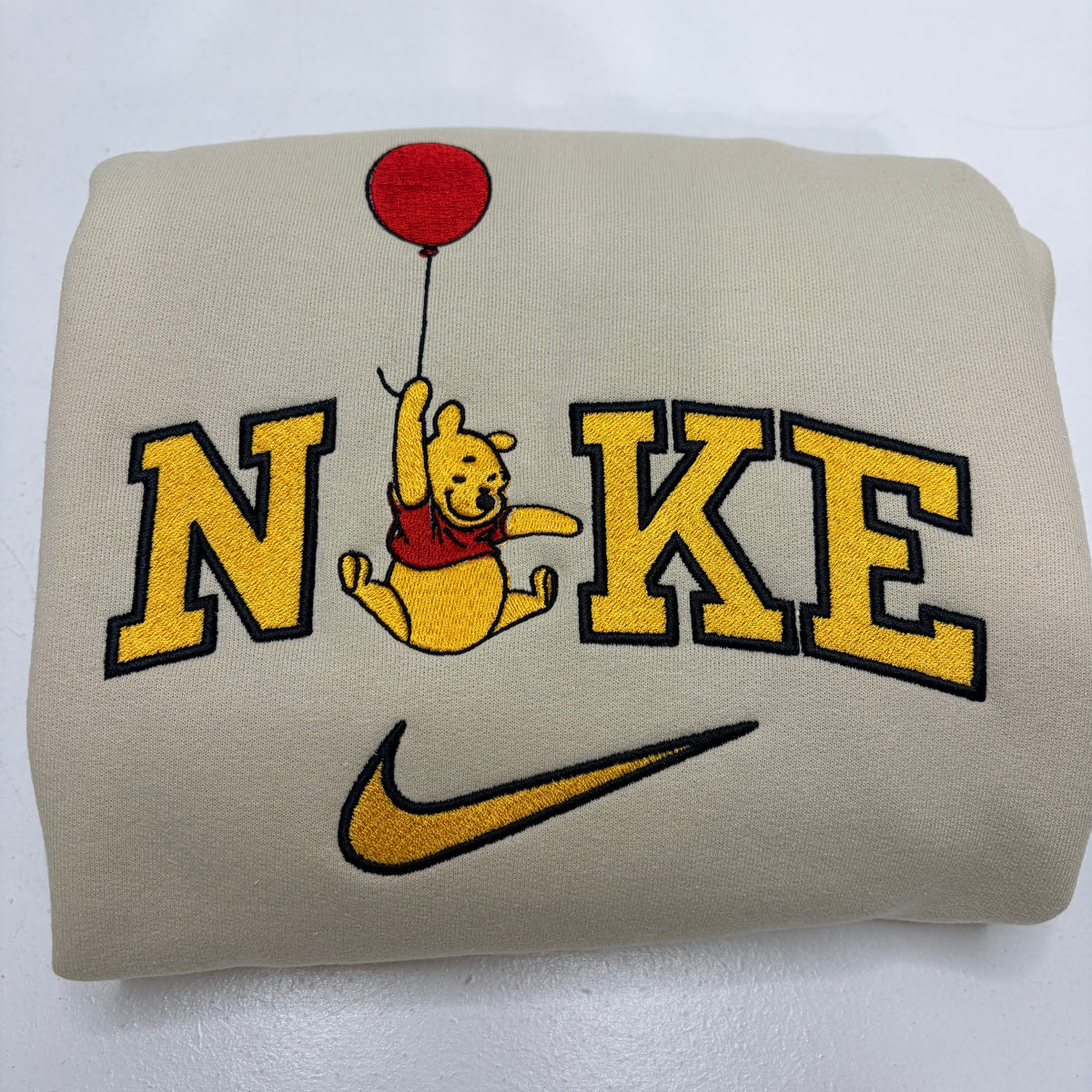 A folded beige sweatshirt featuring a large embroidered design with the word 'NIKE' in yellow varsity letters, Winnie the Pooh holding a red balloon, and a yellow Nike swoosh below.