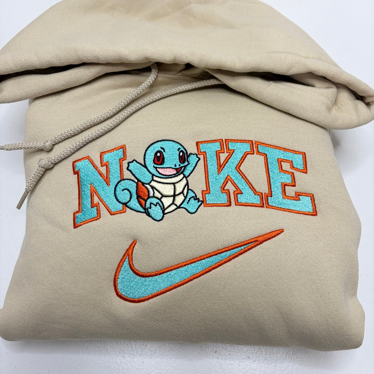 A beige hoodie featuring custom embroidery with the word 'NIKE' in light blue with orange outlines, a Squirtle character in the center, and a matching light blue and orange Nike swoosh below. The hoodie is shown folded on a white background.