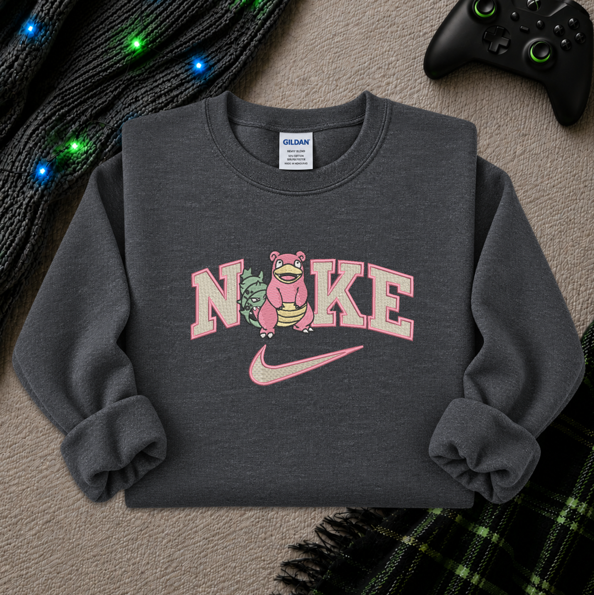 A dark gray crewneck sweatshirt featuring a pink and white graphic that reads 'NIKE' with a pink creature replacing the 'O' and a pink swoosh underneath. The sweatshirt is styled in a flat lay on a textured beige carpet with a black game controller, a black knit blanket with colorful lights, and a green plaid blanket.