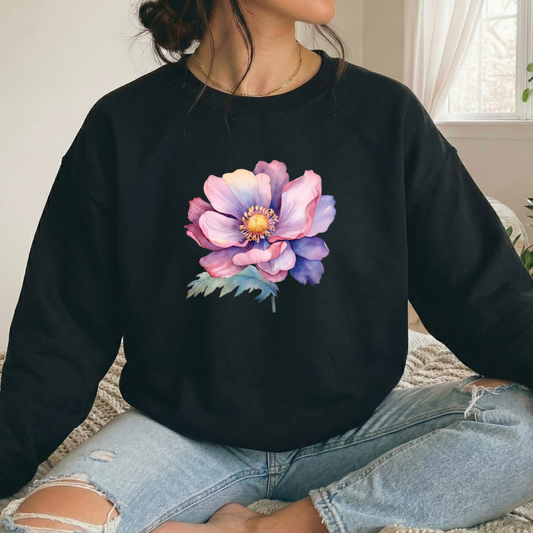 A person sitting cross-legged indoors wearing a black crewneck sweatshirt with a large colorful floral print on the front, paired with light blue ripped jeans.
