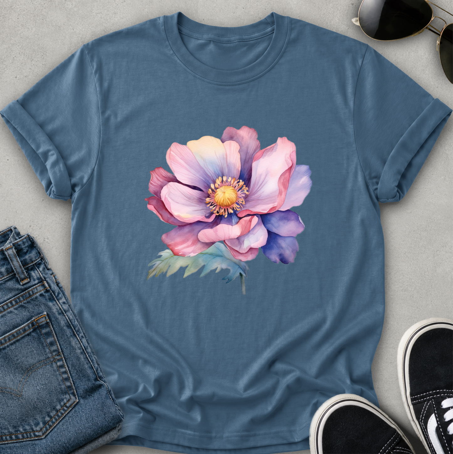 A slate blue short-sleeve crewneck t-shirt featuring a large watercolor-style graphic of a pink and purple peony flower on the chest, displayed flat against a light gray background.
