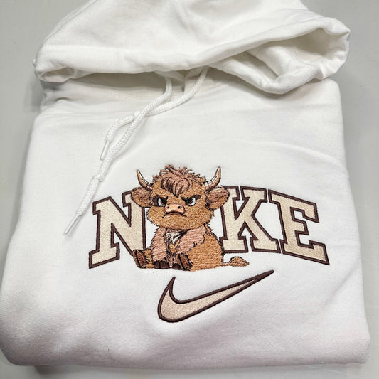 A folded white hoodie featuring an embroidered cartoon Highland cow mascot sitting in front of the word 'NORK' in cream varsity letters with brown outlines, and a brown Nike swoosh below.
