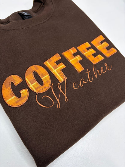 A folded brown crewneck sweatshirt featuring the word 'COFFEE' in a textured orange and yellow plaid font, with 'Wather' written in orange cursive script underneath, displayed on a white background.