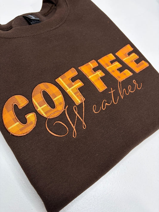 A folded brown crewneck sweatshirt featuring the word 'COFFEE' in a textured orange and yellow plaid font, with 'Wather' written in orange cursive script underneath, displayed on a white background.