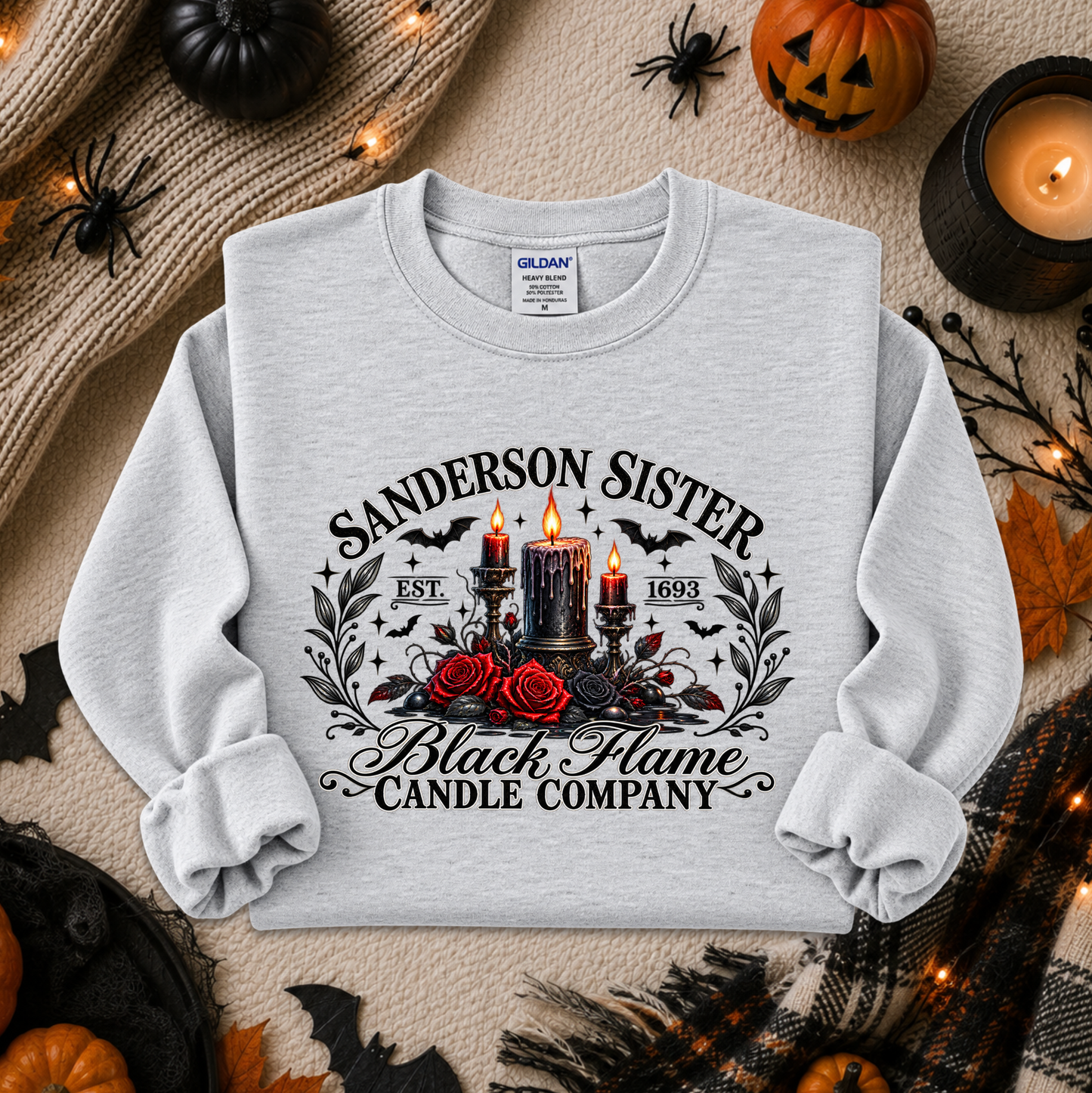 A light heather gray crewneck sweatshirt with a detailed black, red, and gold Halloween graphic reading 'Sanderson Sister Black Flame Candle Company' with candles, roses, bats, and spiders, presented in a flat-lay holiday setting.
