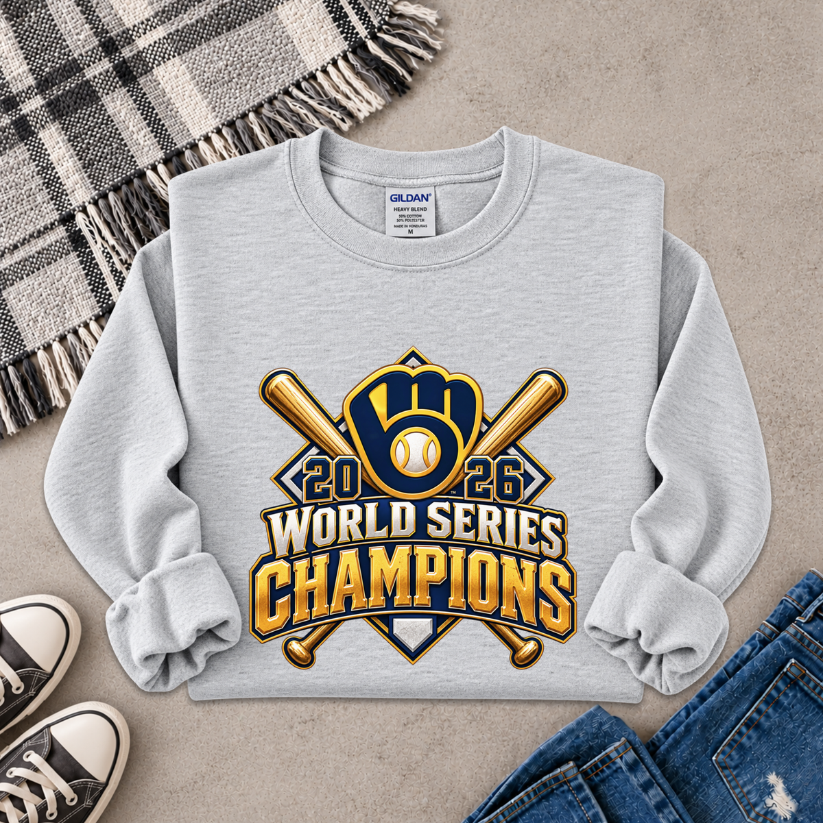 A light gray long-sleeve sweatshirt laid flat on a textured beige surface, featuring a large 2026 World Series Champions baseball graphic in gold, navy, and white. The sweatshirt is styled with a plaid blanket, black sneakers, and blue jeans.