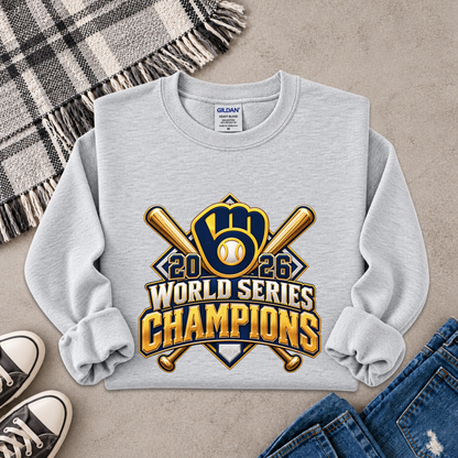 A light gray long-sleeve sweatshirt laid flat on a textured beige surface, featuring a large 2026 World Series Champions baseball graphic in gold, navy, and white. The sweatshirt is styled with a plaid blanket, black sneakers, and blue jeans.