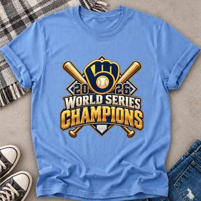 A light blue short-sleeve t-shirt laid flat on a gray surface, featuring a large gold and navy baseball graphic that reads '2026 World Series Champions' with crossed bats and a glove. The shirt is styled with a plaid blanket, black sneakers, and blue jeans.