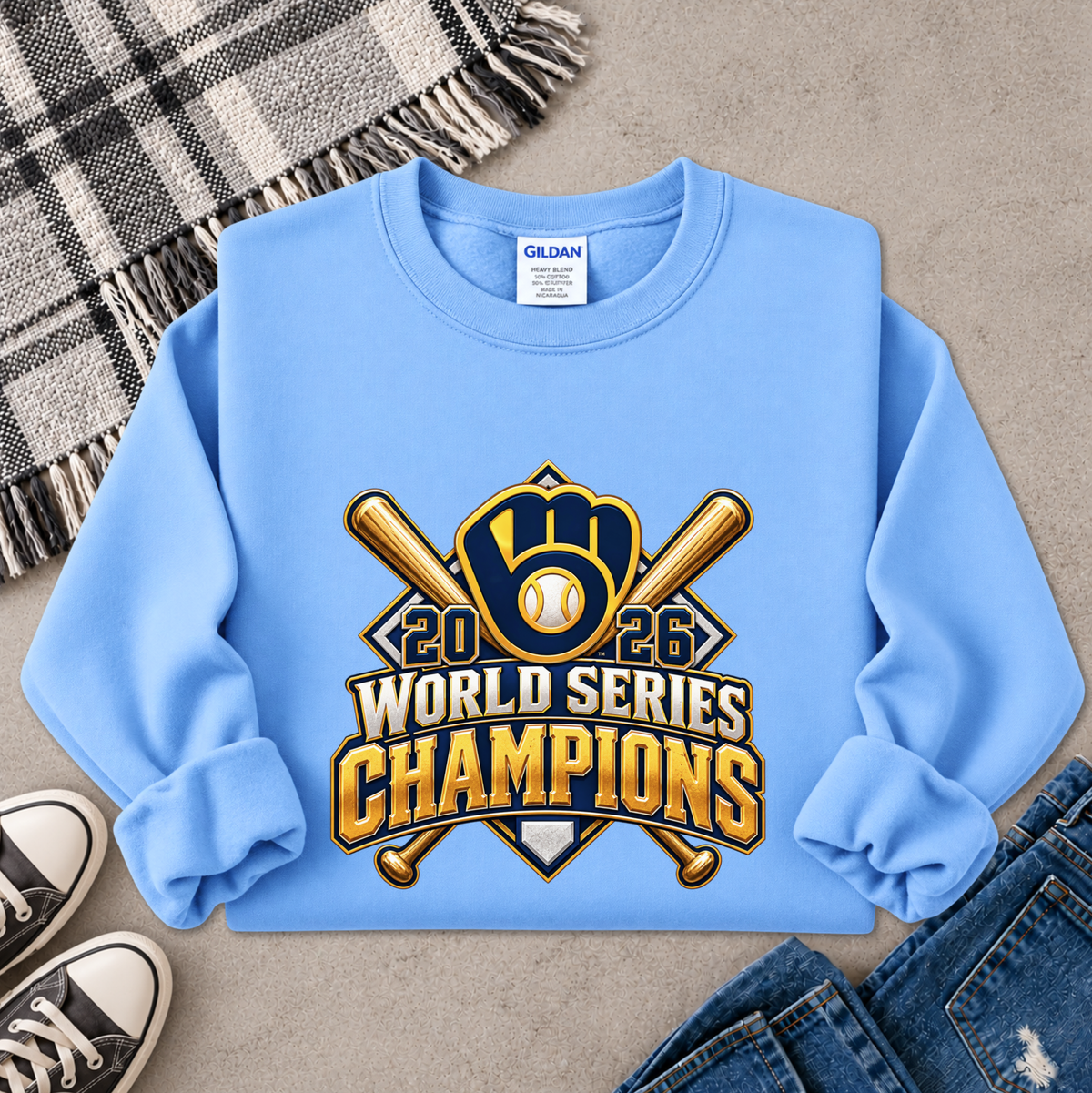 A light blue crewneck sweatshirt laid flat on a gray surface, featuring a large '2026 World Series Champions' graphic with crossed bats, a baseball, and a glove in gold, navy, and white. The sweatshirt is styled with a plaid blanket, black sneakers, and blue jeans.