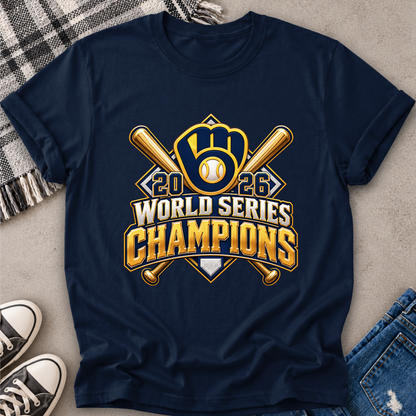 A navy blue short-sleeve t-shirt with a large gold and white '2026 World Series Champions' graphic featuring a baseball glove and crossed bats, styled in a flat lay with jeans, sneakers, and a plaid blanket on a concrete floor.