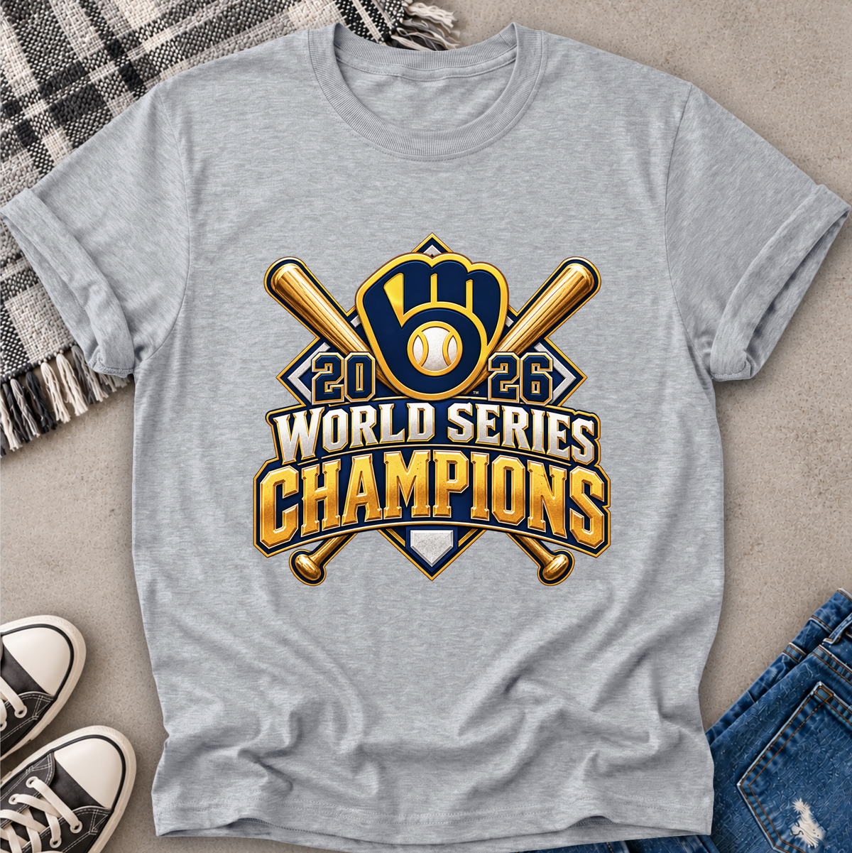 A heather gray short-sleeve t-shirt laid flat on a neutral surface, featuring a large 2026 World Series Champions baseball graphic in gold, navy, and white. The shirt is styled with a plaid blanket, black-and-white sneakers, and blue jeans.