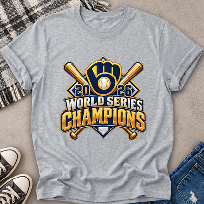 A heather gray short-sleeve t-shirt laid flat on a neutral surface, featuring a large 2026 World Series Champions baseball graphic in gold, navy, and white. The shirt is styled with a plaid blanket, black-and-white sneakers, and blue jeans.
