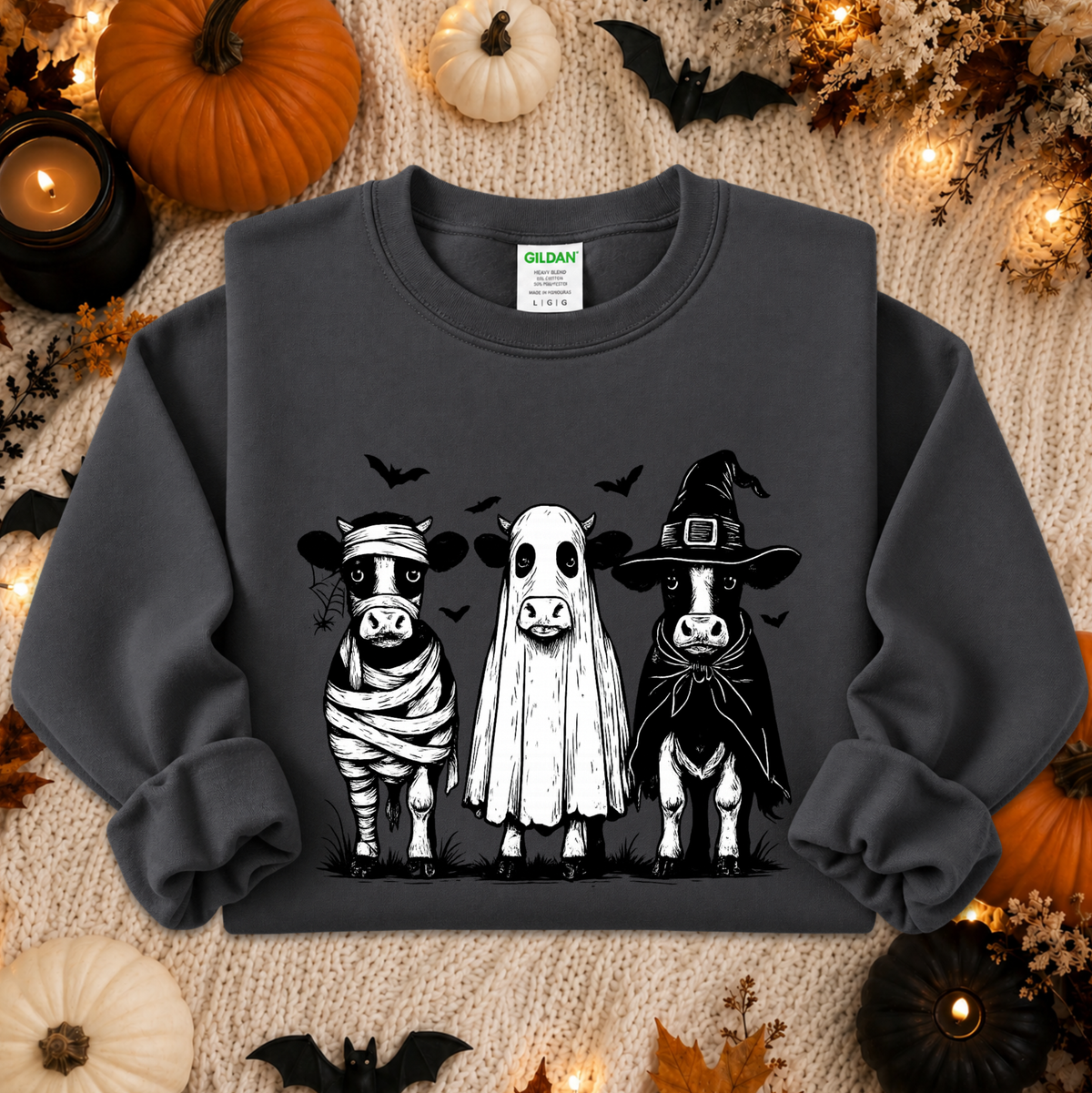 A dark charcoal gray crewneck sweatshirt featuring a graphic of three cute Halloween cows dressed as a mummy, a ghost, and a witch. The sweatshirt is displayed in a flat-lay arrangement on a cozy beige knit blanket, surrounded by seasonal props including pumpkins, candles, and bats.