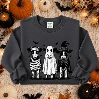 A dark charcoal gray crewneck sweatshirt featuring a graphic of three cute Halloween cows dressed as a mummy, a ghost, and a witch. The sweatshirt is displayed in a flat-lay arrangement on a cozy beige knit blanket, surrounded by seasonal props including pumpkins, candles, and bats.