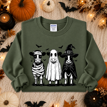 An olive green crewneck sweatshirt featuring a black and white graphic of three cows dressed in Halloween costumes (mummy, ghost, and witch), laid flat on a cozy beige knit blanket surrounded by autumn props like pumpkins, bats, and candles. A Gildan brand tag is visible inside the collar.