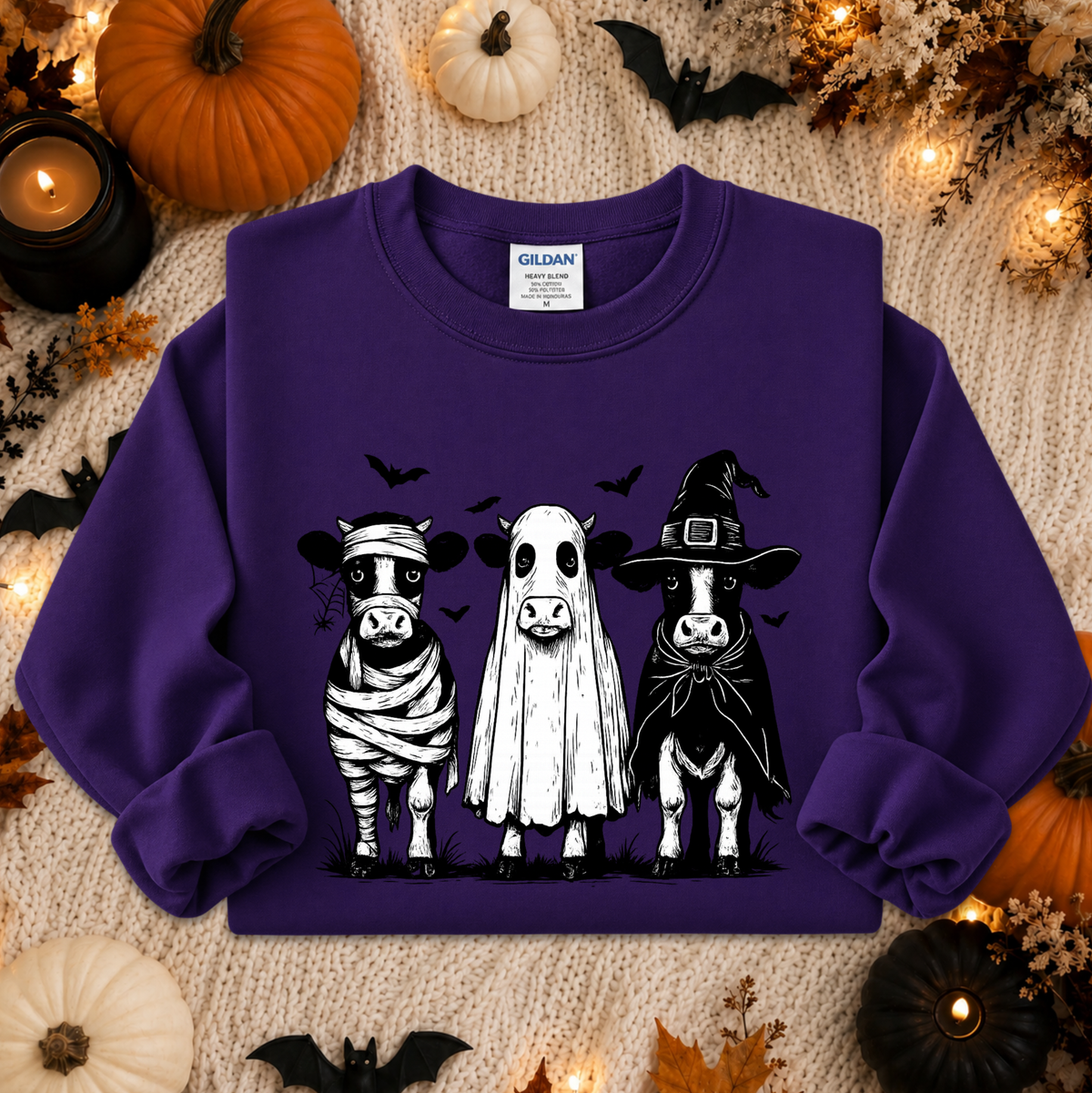 A purple long-sleeve sweatshirt with a graphic of three cute cows dressed in Halloween costumes (bandage, ghost, and witch hat) on a cozy knitted blanket surrounded by pumpkins, candles, and bats.
