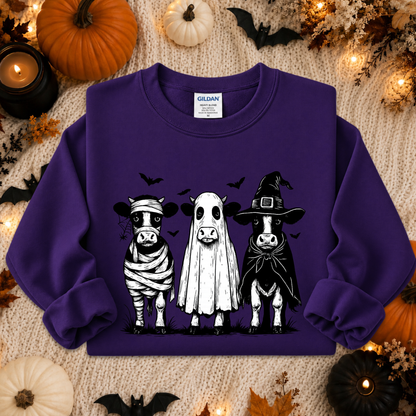 A purple long-sleeve sweatshirt with a graphic of three cute cows dressed in Halloween costumes (bandage, ghost, and witch hat) on a cozy knitted blanket surrounded by pumpkins, candles, and bats.