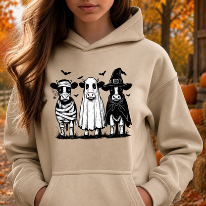 A woman wearing a beige hoodie with a graphic of three cows dressed in Halloween costumes (bandage, ghost, and witch hats) on the front, set against an autumn outdoor background with pumpkins.