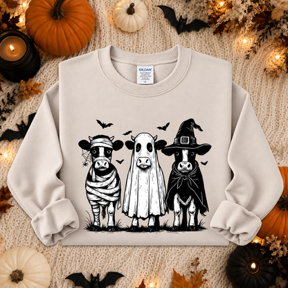 A cream-colored crewneck sweatshirt featuring a black graphic of three cute cows dressed as a mummy, a ghost, and a witch, laid flat on a cozy knit blanket surrounded by Halloween decorations like pumpkins, candles, and bats.