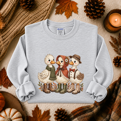 A light gray crewneck sweatshirt featuring a graphic of three cute ducks dressed in autumn clothing, styled in a cozy fall flat-lay with pinecones, leaves, a candle, and a knitted scarf.