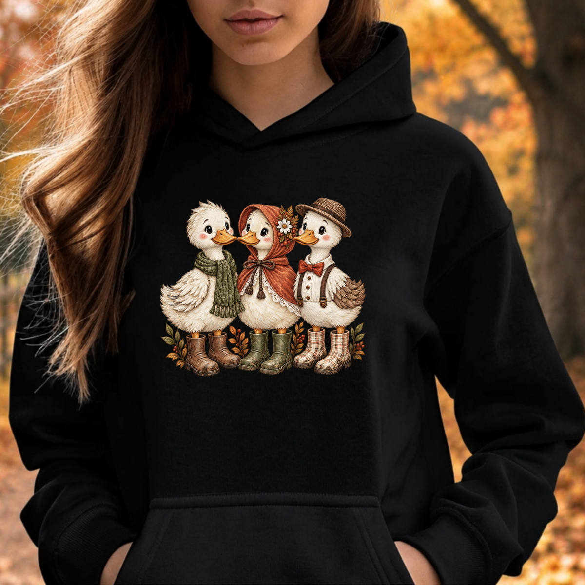 A woman wearing a black pullover hoodie with a graphic of three cute ducks dressed in autumn clothing, set against a blurred outdoor autumn background.