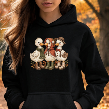 A woman wearing a black pullover hoodie with a graphic of three cute ducks dressed in autumn clothing, set against a blurred outdoor autumn background.