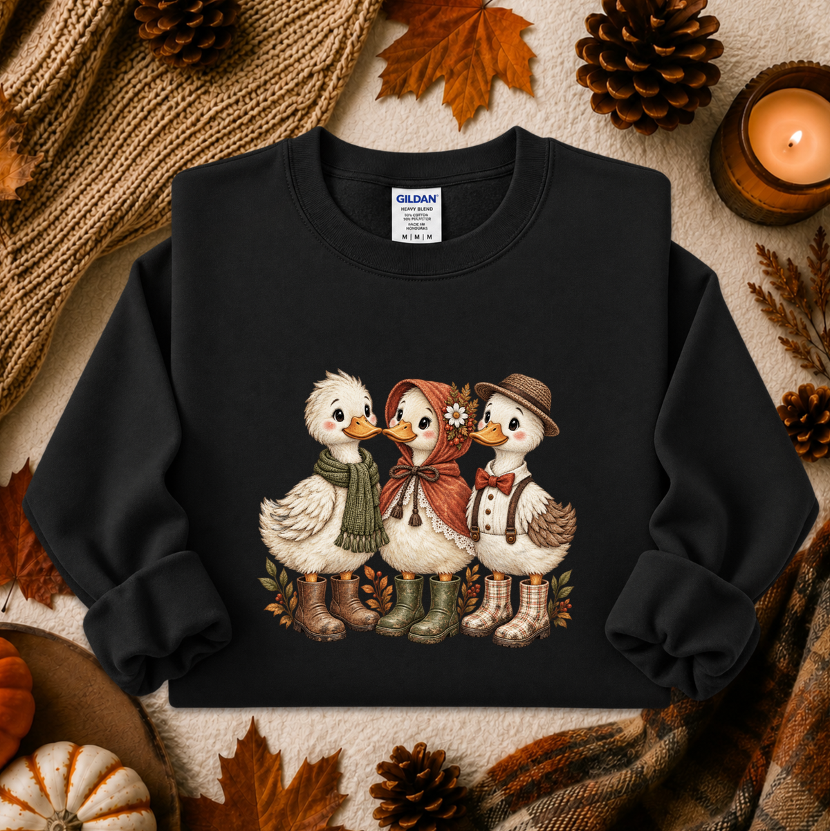 A black crewneck sweatshirt featuring a graphic of three cute ducks dressed in autumn clothing, styled in a cozy fall flat-lay with pinecones, leaves, a candle, and a knit blanket.